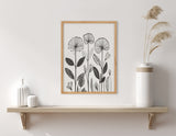 Black and white linocut-style botanical print with tall flowers and varied leaf shapes in a light wood frame above a simple wooden shelf with white ceramic vases and tall grasses.