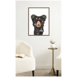 Framed canvas print of a textured black bear portrait with a peach-colored snout and green ears on a white background, displayed above a small brass side table and cream chairs.