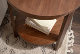 Round brown wood side table with two tiers, placed on a textured beige and brown patterned rug, holding a stack of white books on the lower shelf.