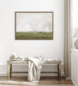 Framed landscape painting with muted green fields under a cloudy, pale sky, hanging above a wooden bench with a draped light cloth and an open book in a neutral room.