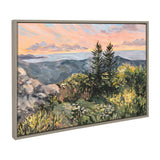 Framed canvas painting of a mountain landscape at sunrise with pine trees, rocky foreground, and a pastel sky of orange and pink hues.