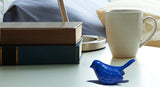Small blue bird figurine with textured detailing placed on a white surface next to a stack of books, a beige patterned mug, and a metallic desk lamp base.