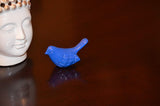 Small glossy blue bird figurine with detailed wing texture placed on a dark polished surface next to a white Buddha head sculpture base.