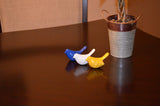 Three small ceramic bird figurines in blue, white, and yellow arranged in a row on a dark polished surface next to a small potted plant in a metal container.