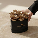 Heart-shaped black box with six preserved gold roses, hand touching the side, placed on beige marble surface with soft natural lighting.