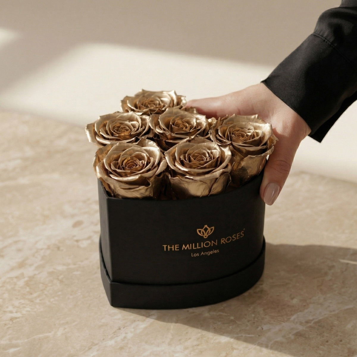Heart-shaped black box with six preserved gold roses, hand touching the side, placed on beige marble surface with soft natural lighting.