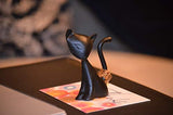 Small black cat figurine with a curved tail, sitting on a colorful card on a dark surface with blurred background details.