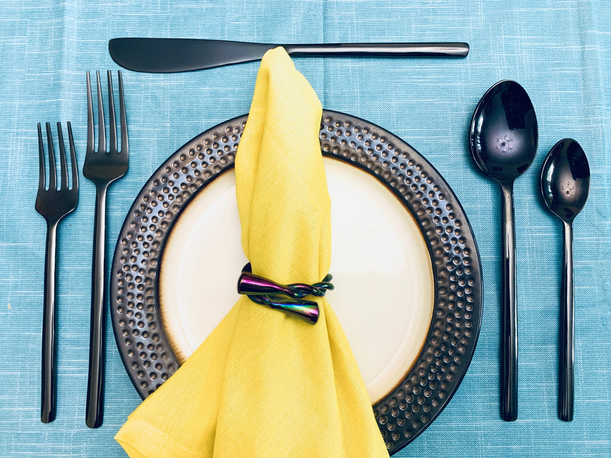 Matte black flatware including knife, forks, and spoons arranged around a cream plate with a hammered dark metal rim, accented by a yellow napkin with a multicolor ring.