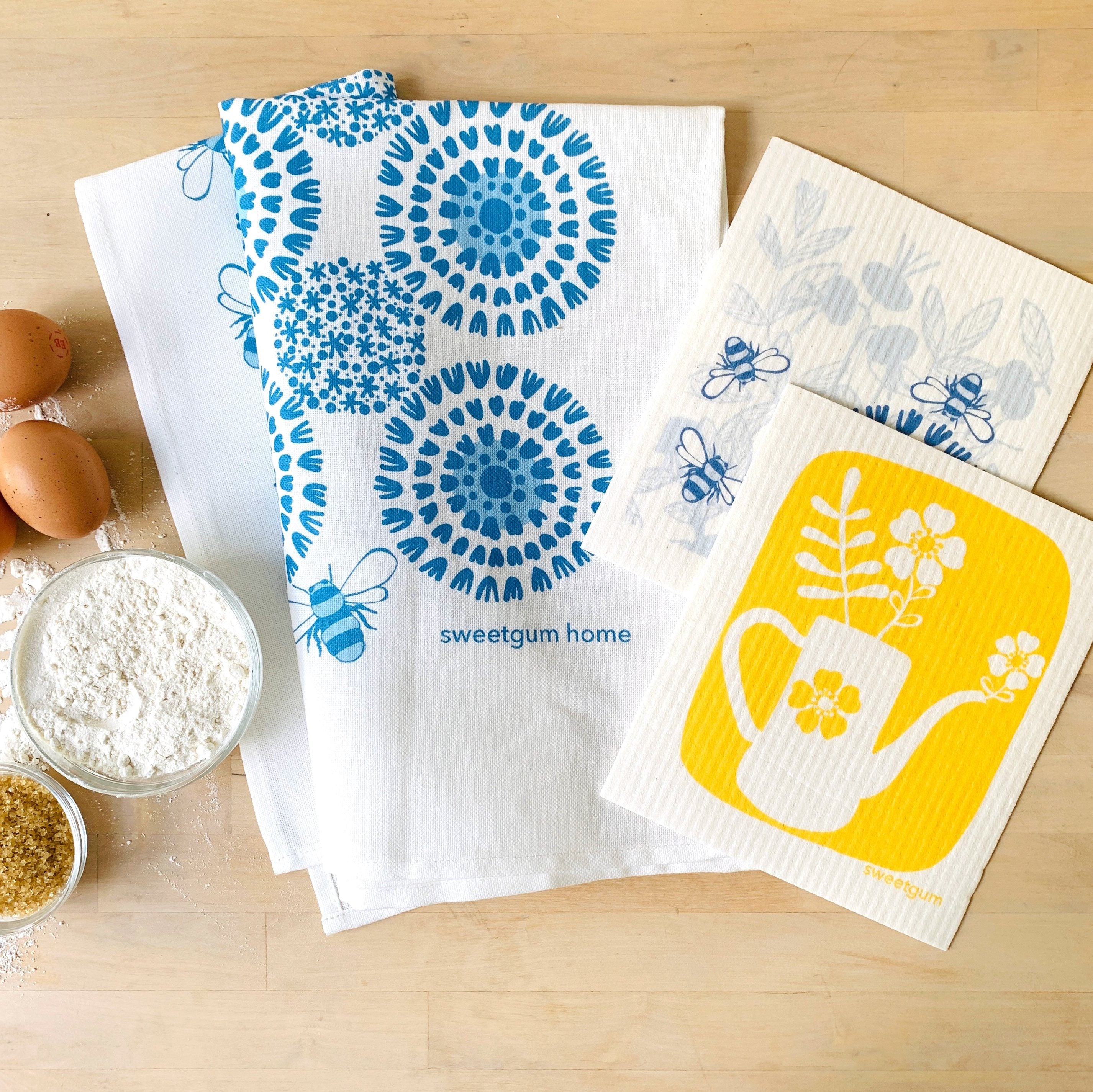 Bloom Tea Towel with yellow teapot and two Swedish dishcloths in blue and yellow designs laid flat on surface