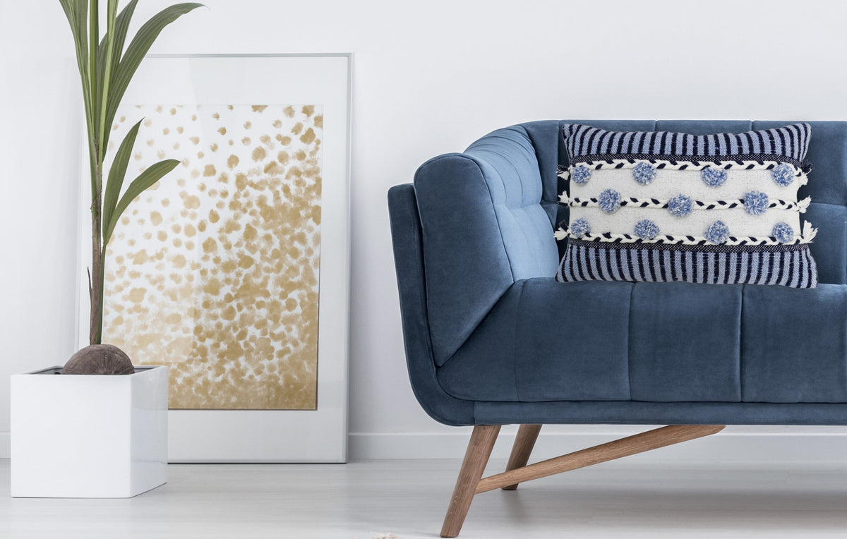 Blue velvet couch with angled wooden legs featuring a striped throw pillow decorated with blue pom-pom braids and cream tassels, next to a potted tall green plant and framed gold speckled art.