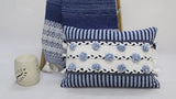 Rectangular striped pillow with blue and white horizontal stripes, braided black and white bands, and blue pom-poms, placed in front of a patterned blue throw and beige pottery.