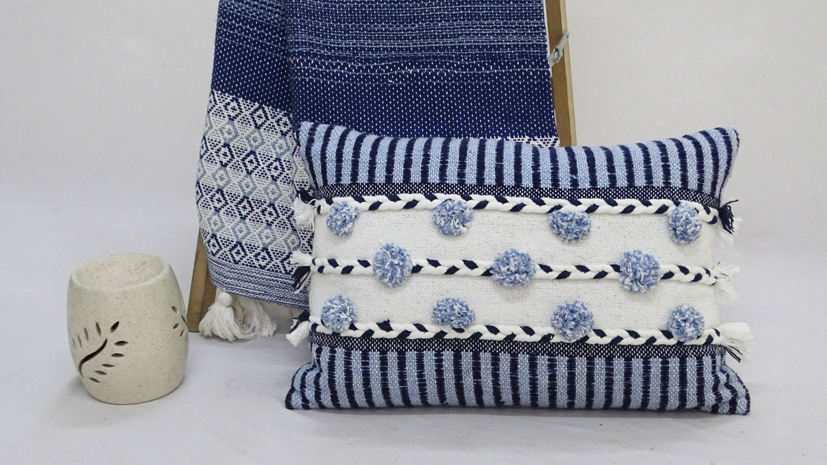 Rectangular striped pillow with blue and white horizontal stripes, braided black and white bands, and blue pom-poms, placed in front of a patterned blue throw and beige pottery.