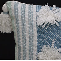 Close-up of a woven accent pillow with vertical blue and white stripes, featuring large white pom-poms and a white tassel on the corner.
