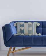 Rectangular pillow with blue and cream stripes, featuring large cream pom-poms and tassels on all corners, resting on a tufted blue velvet couch with wooden legs.