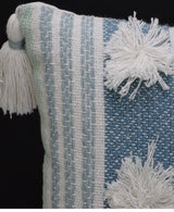 Close-up of a woven accent pillow with vertical blue and white stripes, featuring large white yarn pompoms and a thick tassel on the corner against a dark background.