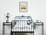 Black metal-framed single bed with white and light blue bedding, flanked by two black side tables holding a plant and a clock, beneath a framed canvas of a yellow van on a desert road.