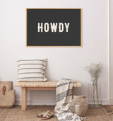 Black canvas framed in wood with the bold word 'HOWDY' in cream letters, hung above a simple wooden bench with striped cushion and throw in a neutral-toned room.