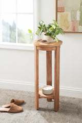 Round wooden side table with a live plant and sunglasses on top, featuring three vertical legs and a lower shelf with a white candle, placed near a window.