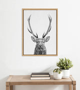 Black and white framed portrait of a red deer with large antlers centered on a white wall above a wooden table with books and potted plants.