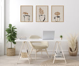 Minimalist white desk with beige chair beneath three framed botanical line art prints featuring vases with brown leaves, flanked by woven baskets holding a green plant and pampas grass.