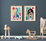 Two framed Sylvie animal-themed canvas prints by Rachel Lee featuring colorful stylized giraffes and elephants with abstract plants on a dark gray wall above children's furniture.