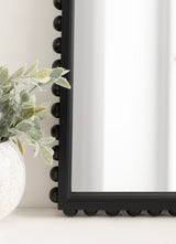 Close-up of a black arched wall mirror frame with rounded bead details along the edge, beside a white vase with soft green leaves on a light surface.