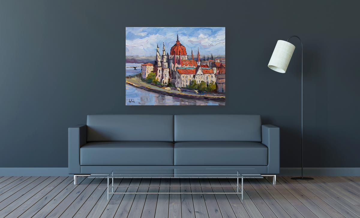 Painting of Budapest's cityscape with red-domed building and spires along a river, hung above a modern gray sofa and next to a curved floor lamp on gray wall.
