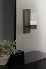 Rectangular dark metal wall sconce with vertical ridges and cylindrical clear glass shade holding a white candle, mounted on light gray wall above black furniture.