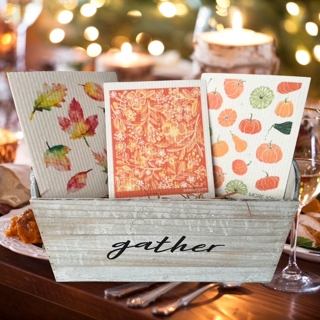 Bundle of three Swedish dishcloths with prints of autumn leaves, orange flowers, pumpkins, and gourds, arranged on a neutral fabric background.