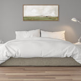Horizontal framed canvas above bed showing muted green and beige landscape with cloudy sky, hanging on gray wall behind white bedding and gray upholstered bed frame.