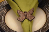 Antique bronze butterfly-shaped napkin ring secured around a folded olive green napkin on a white plate with a black beaded rim.