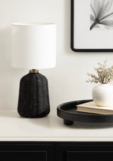Table lamp with a cylindrical white fabric shade and a textured black woven rattan base topped with a brass accent, placed on a white surface beside a black tray and a white vase.