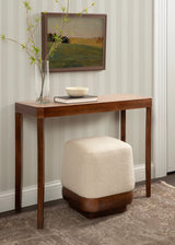 Minimalist wooden console table with gently curved legs above a square cream fabric ottoman featuring a rounded wooden base, set against striped wallpaper and a landscape painting.