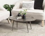 Round gray concrete coffee table with irregular edge and four angled legs, decorated with two black vases with green foliage and two stacked books, in a neutral-toned living room.