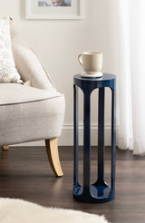 Tall, cylindrical navy blue metal side table with vertical cutouts, standing on dark hardwood floor next to a light beige sofa with wooden legs and a patterned pillow.