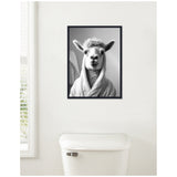 Black and white portrait of a llama wearing a bathrobe, framed and hung on a white bathroom wall above a white toilet tank, with blinds partially visible to the left.