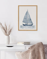 Minimalist framed print of a blue sailboat with striped sails above textured blue waves, hanging on a white wall above a desk with a vase of pampas grass and stacked books.