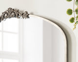 Curved arch mirror with distressed white frame and ornate silver floral carving at the top center, reflecting a window and some greenery.