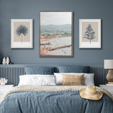 Bedroom with blue-gray walls featuring three framed artworks: a central cityscape with mountains, flanked by minimalist dark blue botanical prints on beige backgrounds.