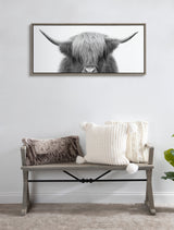 Framed grayscale canvas of a Highland cow’s head with long shaggy hair and wide horns, hung above a wooden bench with textured beige and brown pillows.