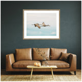 Framed wall art above a brown leather sofa, depicting two cranes flying against a pale blue sky with soft clouds, in a neutral-toned contemporary living room.