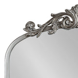 Top corner of an arched mirror with a silver-gray decorative frame featuring ornate scrolls and leaf patterns with a slightly aged finish.