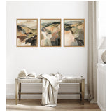 Three framed abstract landscape paintings with soft earthy tones and muted greens hanging in a row above a wooden bench with a draped light beige cloth and an open book.