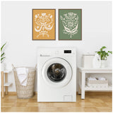 Two framed botanical prints with white floral designs on mustard yellow and green backgrounds hanging side by side above a white front-load washing machine in a laundry room.
