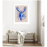 Framed artwork of a blue lobster against a beige background hanging on a white wall above a wooden bench with an open book and light-colored fabric draped over it