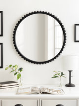 Round wall mirror with a black frame featuring evenly spaced circular accents around its edge, hanging above a white console with plants, books, and a lamp.