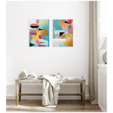 Two framed abstract canvas artworks with geometric shapes in pastel pink, blue, yellow, black, and orange hanging side by side on a white wall above a woven bench with a draped beige cloth.