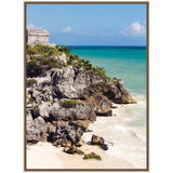 Rocky limestone cliffs with green vegetation overlooking turquoise sea and white sandy beach under blue sky, framed in ornate wood.