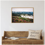 Framed canvas print of a mountain landscape showing snow-capped peaks under cloudy sky, with green meadows and purple wildflowers in the foreground above a wooden bench.