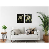 Two framed floral canvas artworks with dark backgrounds featuring white and yellow flowers, hung side by side above a white sofa with patterned cushions in a minimalist living room.